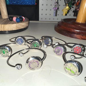 Puede incluir: Una colección de brazaletes de metal plateado, cada uno con un centro circular tipo relicario con un diseño recortado que revela inserciones de colores. Los extremos de cada brazalete están terminados con pequeños cristales redondos y claros.
