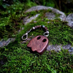 May include: A dark brown wooden ghost-shaped charm featuring two circular cutouts for eyes. The charm is attached to a metal lobster clasp by a short beaded chain consisting of small dark brown beads and silver-toned metal spacers.