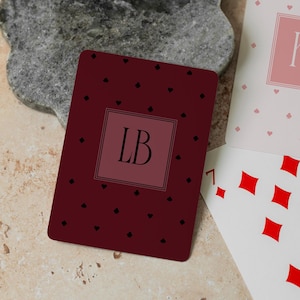 May include: A dark maroon playing card featuring a central square with the letters LB, surrounded by a pattern of small black suit symbols. Beside it, a white playing card displays the letters KR in a pink square and a seven of diamonds card.