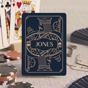 May include: A dark blue playing card featuring a gold-colored line art illustration of a king figure and the name JONES printed in the center. The card is displayed alongside standard playing cards, poker chips, and a metal key.