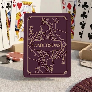 May include: A deck of playing cards featuring a deep purple card back with gold line art of two mirrored figures holding flowers and the text ANDERSONS in a central frame. Standard playing cards including a King of Hearts are visible.