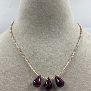 Puede incluir: Un collar que presenta una hilera de pequeñas perlas de semillas color crema. Tres grandes piedras preciosas facetadas en forma de lágrima de color púrpura intenso cuelgan del centro de la hilera, creando un punto focal con sus superficies oscuras.