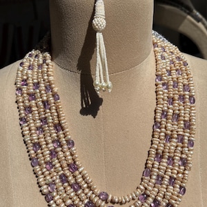 Op de afbeelding: Een meerstrengs ketting met rijen kleine, crèmekleurige zoetwaterparels afgewisseld met gladde, ovaalvormige lichtpaarse amethistkralen. De ketting bevat een zilverkleurig koord met een decoratieve hanger in de vorm van een kralenkwastje.