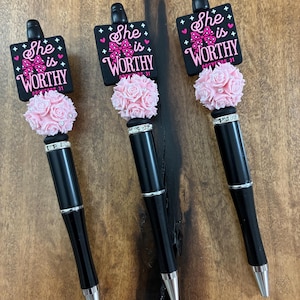 May include: Three black ink pens featuring a square topper with the text "She is WORTHY PROVERBS 31" and a pink bow graphic. Below the topper is a cluster of light pink artificial roses with small silver accents, followed by a silver ring.