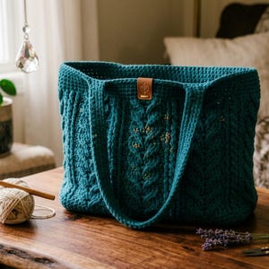 May include: A teal crochet tote bag featuring a textured cable knit pattern and two sturdy shoulder straps. A small rectangular brown leather tag is attached to the top center of the bag. The bag is constructed with thick yarn in a deep blue-green color.