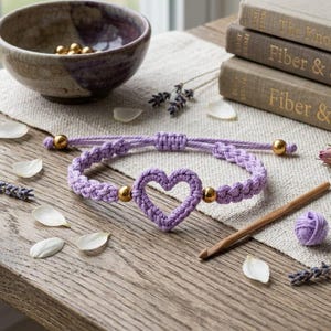 May include: A lavender-colored macrame bracelet featuring a central heart-shaped charm, accented with small gold-toned metal beads on each side. The bracelet includes an adjustable sliding knot closure with gold-toned beads at the ends of the cords.