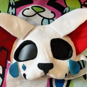 May include: A white plush animal mask featuring large red inner ears, black eye sockets, a black nose, and blue teardrop-shaped accents under the eyes. The mask includes a small red ribbon detail on the left side and an elastic strap for securing.