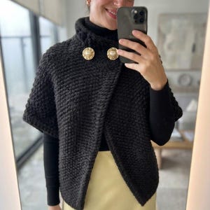 Peut inclure: Une cape en tricot épais gris anthracite foncé avec un col montant et deux gros boutons ronds dorés ornés d'un accent nacré central. Le vêtement présente une texture en laine épaisse et une coupe courte ouverte sur le devant.