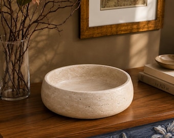 Puede incluir: Un cuenco redondo de piedra travertino beige con una superficie porosa y texturizada. El cuenco tiene un borde grueso y sólido y una base interior lisa. La piedra natural presenta variaciones sutiles de color y textura en su forma.