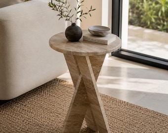 Puede incluir: Una mesa auxiliar redonda de travertino beige con base en forma de cruz. Sobre la mesa hay un jarrón esférico gris oscuro con una rama verde y un pequeño cuenco gris sobre un libro blanco con el texto "LIVE BEAUTIFULLY" en el lomo.