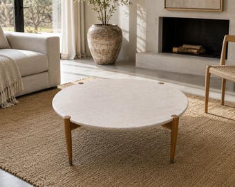 May include: A round coffee table featuring a light beige, speckled stone top with a smooth surface. The table is supported by three tapered, medium-brown wooden legs that connect to the underside of the stone top with visible circular hardware.