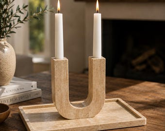 Puede incluir: Un portavelas de travertino beige en forma de U con una textura suave y porosa, que sostiene dos velas cónicas blancas encendidas. El portavelas se asienta sobre una bandeja rectangular de travertino a juego, creando un conjunto de piedra.
