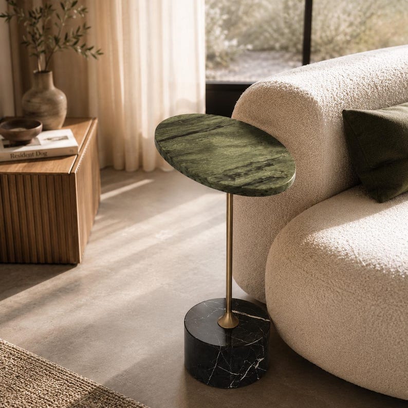 May include: A side table featuring an oval-shaped green marble top, a slender gold-toned metal stem, and a cylindrical black marble base with white veining. The table stands independently with a balanced structure and a polished stone finish.