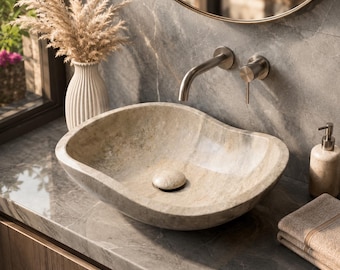 Puede incluir: Un lavabo de piedra beige de forma ovalada con un acabado texturizado natural y una tapa de desagüe de piedra a juego. El lavabo está montado sobre una encimera de mármol gris con un grifo de pared de níquel cepillado y un mando monomando.