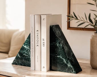 Puede incluir: Dos sujetalibros triangulares hechos de mármol verde oscuro con vetas blancas, colocados a ambos lados de tres libros blancos. Los libros llevan los títulos "Kinfolk" y "at 100" con texto visible en sus lomos, sostenidos por los soportes de piedra.