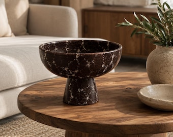 Puede incluir: Un cuenco con pedestal de color marrón oscuro que presenta un patrón de motas blancas en toda su superficie. El cuenco tiene una abertura circular ancha y se asienta sobre una base robusta y cónica, creando una silueta elevada y decorativa.