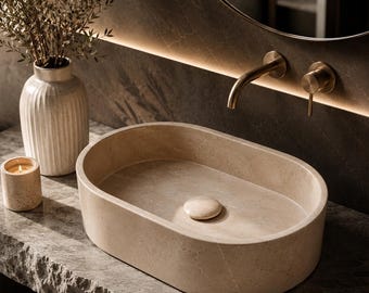 Puede incluir: Un lavabo de piedra beige con forma ovalada y tapa de desagüe a juego. El lavabo está colocado sobre una encimera de piedra gris texturizada junto a una pequeña vela cilíndrica y un jarrón estriado de color blanco roto con follaje seco.