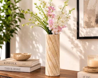 Puede incluir: Un jarrón cilíndrico de piedra beige con una superficie texturizada en espiral, que contiene un ramo de flores delicadas blancas y rosa claro con tallos verdes. El jarrón está en posición vertical, mostrando un acabado poroso natural.