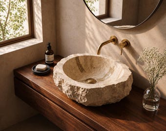 Puede incluir: Un lavabo redondo de piedra natural beige con un exterior rugoso y texturizado y una cubeta interior lisa. Cuenta con un grifo de pared y un desagüe de latón, colocados sobre un mueble de madera oscura junto a una jabonera y un florero.