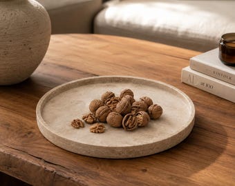 Puede incluir: Una bandeja redonda de piedra beige texturizada que contiene un grupo de nueces marrones enteras y partidas. La bandeja presenta un borde bajo elevado y un acabado de superficie poroso y natural, creando una base neutra.