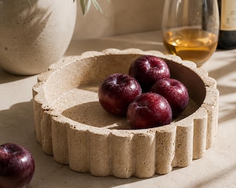 Puede incluir: Un cuenco redondo de piedra beige texturizada con un borde festoneado y acanalado. Dentro del cuenco hay cuatro ciruelas lisas de color rojo oscuro. Una ciruela roja oscura adicional se encuentra sobre la superficie fuera del cuenco.