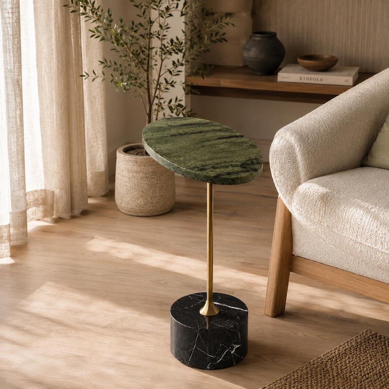 May include: A side table featuring an oval-shaped green marble top supported by a slender gold-toned metal stem and a cylindrical black marble base with white veining, standing as a singular piece of furniture with a polished stone finish.