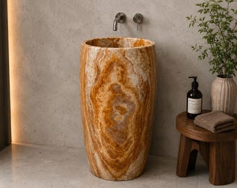 Puede incluir: Un lavabo de pedestal cilíndrico y exento, fabricado en piedra ónix pulida de tonos ámbar y crema con patrones naturales en espiral. La cubeta presenta un borde liso y redondeado, destacando las vetas orgánicas y únicas de la piedra natural.