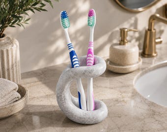 Puede incluir: Un soporte para cepillos de dientes con patrón de mármol gris y diseño escultórico curvo que sostiene dos cepillos. Un cepillo tiene mango azul y blanco con cerdas azules, y el otro tiene mango rosa y blanco con cerdas verdes.