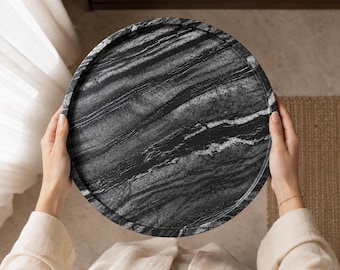 Puede incluir: Una bandeja circular con un patrón de mármol en blanco y negro con vetas lineales distintivas. La bandeja tiene un borde elevado y una superficie lisa y pulida, mostrando una apariencia de piedra natural con vetas oscuras y claras contrastantes.