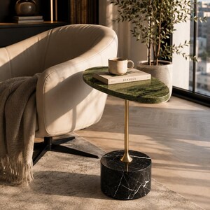 May include: A side table featuring an oval green marble top, a slender brass-toned metal stem, and a cylindrical black marble base with white veining. A beige ceramic mug and a book titled KINFOLK are placed on the table surface.