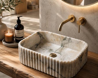 Puede incluir: Un lavabo rectangular de mármol con un diseño exterior acanalado y vetas grises y blancas. El lavabo incluye un tapón de drenaje circular de latón y se combina con un grifo y manija de latón montados en la pared, sobre una superficie de madera.
