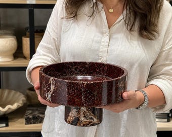 Puede incluir: Un cuenco de pedestal de mármol rojo oscuro y marrón con un acabado liso y pulido y vetas blancas visibles en toda la piedra. El cuenco presenta una cuenca circular ancha apoyada por una base cilíndrica gruesa de la misma piedra veteada.