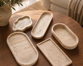 Puede incluir: Una colección de cinco bandejas de piedra travertino beige en varias formas, incluyendo ovaladas, rectangulares y una forma irregular similar a una nube. Cada bandeja presenta una superficie porosa y texturizada con tonos naturales.
