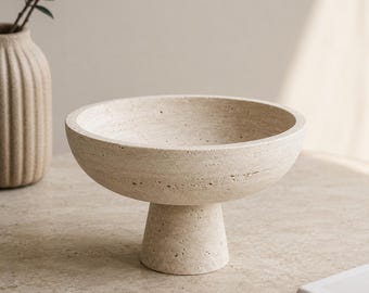Puede incluir: Un cuenco de piedra travertino beige con un recipiente circular ancho y una base de pedestal gruesa y cónica. La textura porosa natural de la piedra es visible en toda la superficie, mostrando un tono neutro y una construcción sólida y firme.