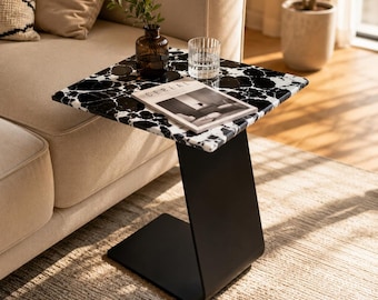 May include: A C-shaped side table featuring a square top with a black and white marbled pattern and a solid matte black metal base. The table holds a clear glass vase with green foliage, a clear drinking glass, and a magazine titled CEREAL.