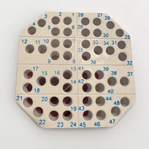 May include: An off-white, octagonal plastic organizer tray featuring a grid of circular holes. The surface is divided into four sections, each containing numbered slots from 1 to 48 in blue ink, designed to hold small items securely in place.