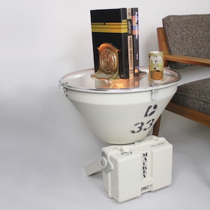 May include: A unique, white, cone-shaped side table with black stenciled numbers and a metal tray top. It holds books, a gold bookend, and a small can. A white box labeled "MACKEY 1967" sits below. A wooden chair is to the right.