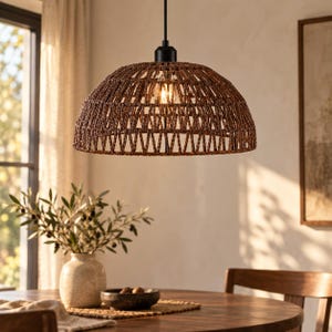 Op de afbeelding: Een koepelvormige hanglamp met een gevlochten bruine rotan kap met een open rasterpatroon. De kap hangt aan een zwart snoer en armatuur en bevat een enkele gloeilamp die een warme gloed door het vlechtwerk verspreidt.