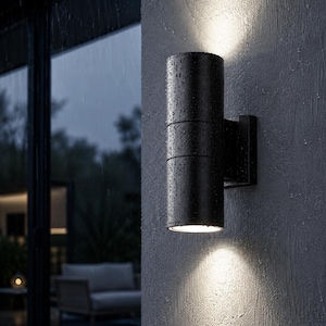 Peut inclure: Une applique murale extérieure cylindrique noire fixée sur un mur gris texturé. Le luminaire présente deux ouvertures lumineuses en haut et en bas, projetant des faisceaux de lumière vifs vers le haut et vers le bas sur la surface du mur.