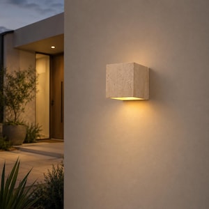 May include: A square wall-mounted light fixture with a textured beige stone finish. The fixture emits a warm, downward-facing glow against a smooth, light-colored exterior wall, creating a soft illumination pattern on the surface below.