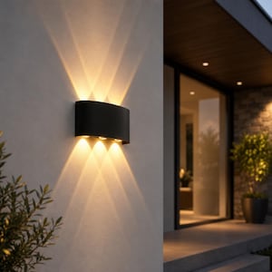 Pode incluir: Uma luminária de parede semicilíndrica em preto mate fixada numa parede exterior de cor clara. A luminária emite feixes de luz quentes e focados tanto para cima como para baixo, criando padrões de estrela distintos na parede.