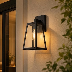 Op de afbeelding: Een matzwarte buitenwandlamp met een trapeziumvormig frame en heldere glazen panelen. Het armatuur is aan een muur bevestigd en bevat een enkele gloeiende lamp in Edison-stijl die een warm licht werpt op het omliggende oppervlak.