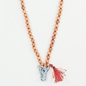 May include: A necklace featuring a long strand of round, light brown wooden beads strung on a dark cord. The necklace includes a central pendant shaped like a gray, textured animal head and a small, vibrant red tassel attached to the side.