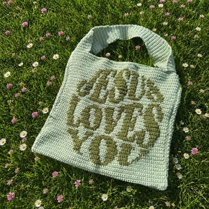 May include: A light sage green crochet tote bag featuring a single shoulder strap and the dark olive green text "JESUS LOVES YOU" centered on the front. The bag is constructed with a dense, uniform stitch pattern and has a square shape.