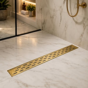 May include: A rectangular, brushed gold linear shower drain featuring a series of horizontal, slotted cutouts. The drain is installed flush within a white marble floor tile surface, matching the surrounding marble wall tiles and gold fixtures.