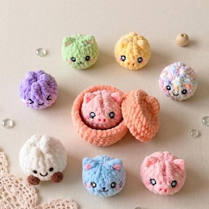 May include: A collection of small, round, crocheted plush toys featuring various pastel colors including light green, yellow, purple, white, blue, and pink. One pink plush sits inside a small, round, peach-colored crocheted basket with a lid.