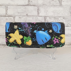 May include: Black wallet with a colorful cartoon design. The wallet features a pattern of yellow stars, blue creatures, purple flowers, and other whimsical elements against a black background with paint splatters. The wallet is displayed on a clear stand.