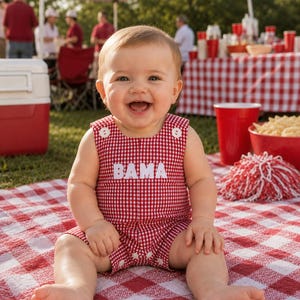 May include: A red and white gingham romper featuring the word BAMA in white block letters on the chest. The sleeveless garment has a round neckline with two white buttons on the shoulder straps and a snap closure at the bottom for easy access.