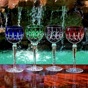 May include: Four crystal wine glasses featuring a diamond-cut pattern on the bowl and a textured, faceted stem. The glasses are colored in deep blue, emerald green, clear, and ruby red, each resting on a clear, circular, starburst-patterned base.