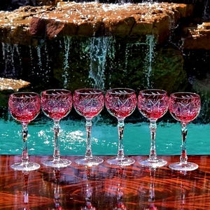 Puede incluir: Un juego de seis copas de vino con tallo que presentan un tono rojo arándano intenso y complejos patrones de cristal tallado. Cada copa tiene un tallo transparente facetado y una base circular, con grabados detallados de hojas.